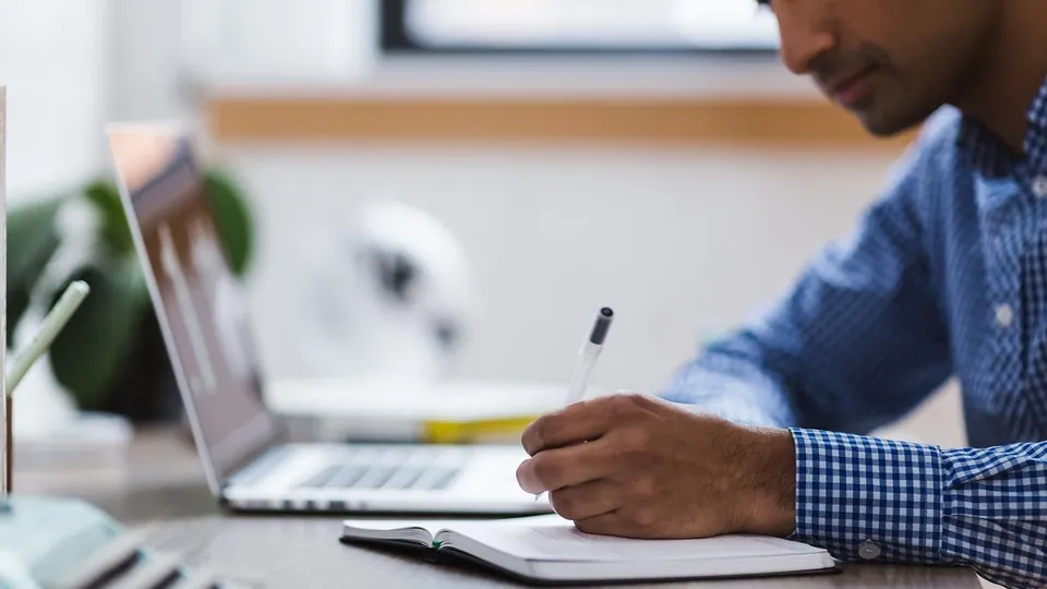 Person am Schreibtisch, die neben dem aufgeklappten Laptop Notizen durchgeht